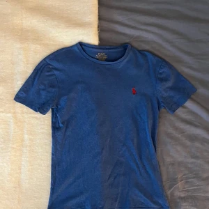 Blå t-shirt från Ralph Lauren - Säljer en blå t-shirt från Ralph Lauren med röd logga på bröstet. T-shirten har en custom slim fit och är kortärmad. Perfekt för en stilren och avslappnad look.