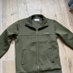 Olivgrön jacka från Les Deux - Snygg olivgrön jacka från Les Deux med dragkedja framtill. Jackan har en stilren design med ribbad krage och långa ärmar. Perfekt för en casual look. I storlek S