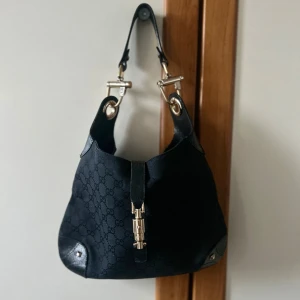 Gucci Jackie väska  - Snygg svart axelväska med monogrammönster och eleganta metalldetaljer. Väskan har en stilren design med en praktisk axelrem och dekorativa spännen. Perfekt för att ge din outfit en lyxig touch. Pris kan diskuteras vid snabb affär.