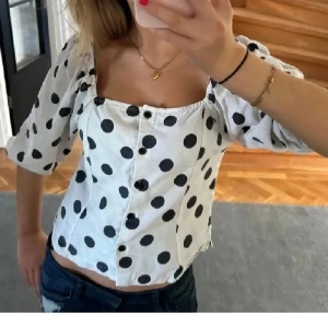 Vit prickig blus - Säljer en söt vit blus med stora svarta prickar och knappar framtill. Blusen har korta puffärmar och en fyrkantig urringning. Perfekt för en trendig look!
