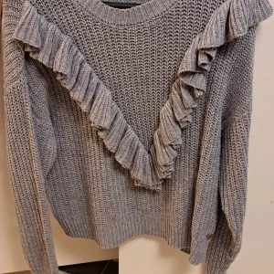Grå stickad tröja med volangdetaljer - Säljer en snygg grå stickad tröja från Ü med volangdetaljer i V-form framtill. Tröjan har långa ärmar och en bekväm passform, perfekt för kyliga dagar. Köpt för några år sedan använts endå mycket skulle jag säga. Fortfarande i bra skick, finns inga hål! 