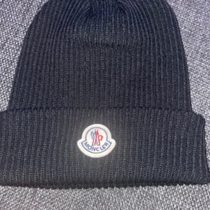 Svart ribbad mössa från Moncler - Snygg svart ribbad mössa från Moncler med deras ikoniska logotyp framtill. Perfekt för att hålla värmen under kyliga dagar. Stilren design som passar till det mesta.