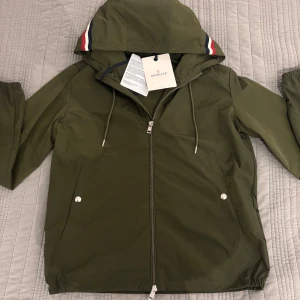 Moncler Grimpeurs Windbreaker - Säljer en nästan oanvänd snygg olivgrön vindjacka från Moncler med huva och dragkedja. Jackan har två fickor med tryckknappar och Moncler-logga på bröstet. Perfekt för blåsiga dagar!