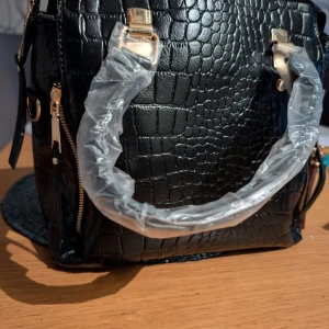 Svart axelväska med krokodilmönster - Snygg svart axelväska i syntet med krokodilmönster och guldfärgade detaljer. Väskan har en praktisk dragkedja och en dekorativ tofs. Perfekt för att ge din outfit en trendig touch.