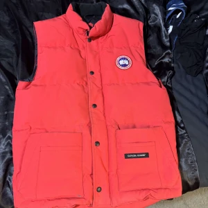 Canada goose väst - Säljer en snygg röd dunväst från Canada Goose med knappar och två stora fickor framtill. Västen har en broderad logotyp på bröstet och är perfekt för kyligare dagar. Passar bra för både höst och vinter. (ÄKTA)!!