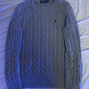 Grå stickad tröja från Ralph Lauren - Säljer en stilren grå stickad tröja från Ralph Lauren med klassiskt flätmönster. Tröjan har långa ärmar och en broderad logotyp på bröstet. Perfekt för kyliga dagar!