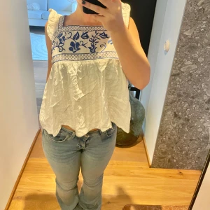 Vit blus med blått broderi och svagt randigt mönster - Vit blus med blått broderi och volangdetaljer och en rosett knytning i ryggen. Svagt randig med små prickar. Super söt i storlek XS