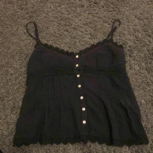 Svart linne från Zara - Säljer ett svart linne från Zara med justerbara axelband och knappar framtill. Linnet har fina spetsdetaljer längs kanterna och en luftig passform. Perfekt för varma dagar eller som ett lager under en kavaj.