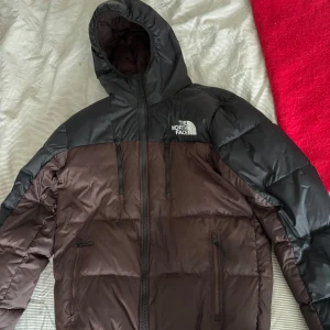 Brun och svart dunjacka från The North Face - Säljer en snygg brun och svart dunjacka från The North Face. Jackan har en huva och dragkedja framtill samt flera praktiska fickor med dragkedjor. Perfekt för kyliga dagar.  (KAN GÅ NER I PRIS VID SNABB AFFÄR)