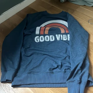 Blå sweatshirt med tryck - Säljer en blå sweatshirt med ett retroinspirerat tryck av en regnbåge och texten 'GOOD VIBES' på framsidan. Tröjan har långa ärmar och ribbade muddar vid ärmslut och nederkant. Perfekt för en avslappnad stil.