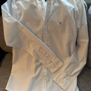 Vit oxfordskjorta från Gant - Vit shield oxford shirt från Gant.  Snygg vit skjorta från Gant med broderad logga på bröstet. Skjortan har långa ärmar och knappar framtill. Perfekt för en stilren look. Anvånd fåtal gånger. Köpt på Kids Brandstore. Ord pros 799.