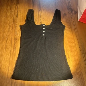 Svart ribbad linnetopp med knappar - Säljer en svart ribbad linnetopp med knappar framtill. Toppen är ärmlös och har en enkel design som passar perfekt till vardags. Perfekt för varma dagar eller som ett basplagg i garderoben.