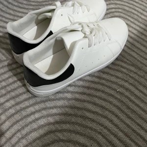 Vita sneakers med svarta detaljer - Snygga vita sneakers med svarta detaljer på hälen. Skorna har klassisk snörning och en stilren design som passar perfekt till vardagsoutfiten. Skorna är helt nya använt 1 gång köpt för 170kr