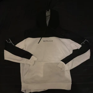 Vit och svart hoodie från Brave Soul - Säljer en stilren vit och svart hoodie från Brave Soul med texten 'MCMLXXII' på bröstet. Hoodien har en klassisk design med långa ärmar och en stor ficka framtill. Perfekt för en avslappnad look.