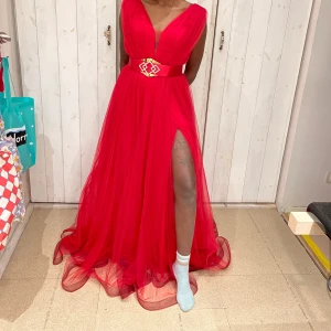 Röd långklänning med slits - Elegant röd långklänning med djup v-ringning och en iögonfallande slits. Klänningen har ett brett bälte med glittrande detaljer i midjan som ger en lyxig touch. Perfekt för speciella tillfällen.