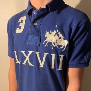 Blå pikétröja från Ralph Lauren - Säljer en blå pikétröja från Polo Ralph Lauren med speciellt tryck. Tröjan har korta ärmar och en klassisk krage med knappar. Perfekt för en stilren look.