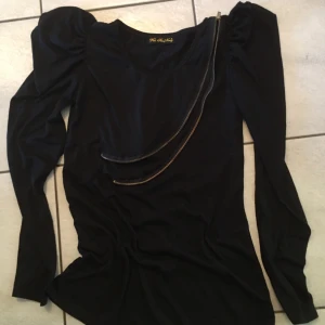 Svart långärmad topp med dragkedja - Snygg svart långärmad topp med unik dragkedjedetalj över bröstet. Perfekt för en trendig look med puffiga axlar som ger extra stil. Passar bra till både jeans och kjol.