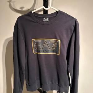 Svart tröja från Emporio Armani EA7 - Snygg svart tröja från Emporio Armani EA7 med ett stort tryck på framsidan. Tröjan har långa ärmar och en rund halsringning. Perfekt för en stilren och avslappnad look.nypris 1100kr