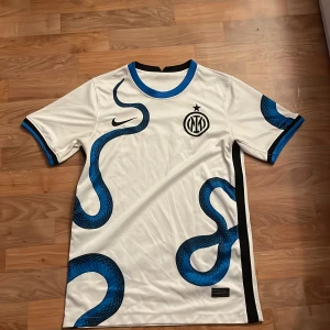 INTER MILAN TRÖJA - Snygg vit fotbollströja med blått ormmönster och svart Inter-logga. Kortärmad design med svarta och blå detaljer vid ärmslut och krage. Perfekt för fans av Inter och fotboll.