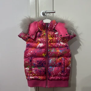 Färgglad dunväst från John Galliano kids - Säljer en dunväst från John Galliano Gazette Pink Down Väst med ett mönster i olika färger. Västen har en avtagbar huva med grå pälsdetaljer och dragkedja framtill.