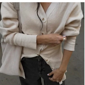 Beige kofta med knappar - Säljer en stilren beige kofta med knappar framtill. Koftan har en v-ringad design och långa ärmar, perfekt för lager-på-lager-stil. Passar till både jeans och kjol för en avslappnad look.