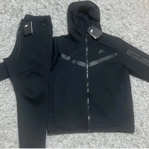 Svart Nike tech fleece  - Säljer en stilren svart hoodie från Nike med dragkedja och huva. Tröjan har en diskret Nike-logga på bröstet och en praktisk ficka med dragkedja på ärmen. Perfekt för en sportig look.