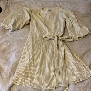 Randig omlottklänning från Adoore - Snygg randig omlottklänning från Adoore i beige och vit. Klänningen har puffiga långärmar och ett knytband i midjan för en smickrande passform. Perfekt för en stilren look.