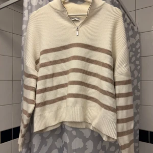 Randig stickad tröja från MNG - Säljer en mysig stickad tröja från MNG i krämvitt med bruna ränder. Tröjan har en dragkedja vid halsen och långa ärmar. Perfekt för kyliga dagar!