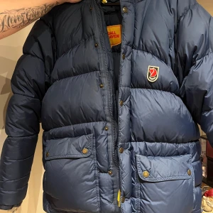 Blå dunjacka från Fjällräven - Säljer en blå dunjacka från Fjällräven med dragkedja och knappar. Jackan har långa ärmar och praktiska fickor framtill. Perfekt för kalla dagar.