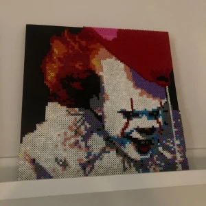 Pennywise tavla. Pärlplatta på canvas tavla.  - En detaljerad pärlplatta föreställande en skrämmande clown med rött hår och vit ansiktsmålning. Tavlan har en mörk bakgrund och är gjord med små, färgglada pärlor som skapar ett pixelartat utseende. Perfekt för den som gillar unika och handgjorda konstverk.              40 x 40 cm