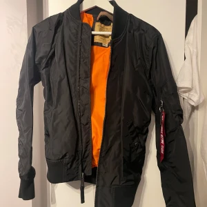 Svart bomberjacka från Alpha Industries - Snygg svart bomberjacka från Alpha Industries med orange foder. Jackan har en dragkedja framtill och en klassisk röd tagg på ärmen. Perfekt för en cool och avslappnad stil.