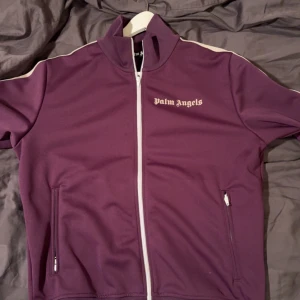 Lila tjocktröja från Palm Angels - Storlek M. Snygg lila track jacket från Palm Angels med vita detaljer och dragkedja. Jackan har långa ärmar och en relativt hög krage. Väldigt ovanlig färg med otroligt bra skick 9,5/10. Köpt 2021 på Kicksnstuff i Göteborg. Använd Max 10 gånger