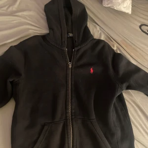 Kofta från Polo Ralph Lauren - Säljer en svart huvtröja med dragkedja från Polo Ralph Lauren. Tröjan har en röd broderad logotyp på bröstet och en praktisk huva. Perfekt för en avslappnad stil.