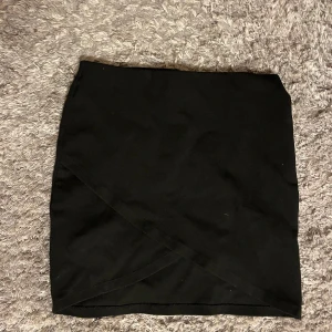 Svart kjol från Gina Tricot - Snygg svart kjol från Gina Tricot i omlott-design. Perfekt för en stilren look. Kjolen är kort och passar bra till olika tillfällen.