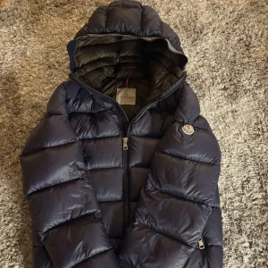Moncler jacka - Säljer en snygg mörkblå dunjacka från Moncler med huva och dragkedja. Jackan har en quiltad design och Moncler-logga på ärmen. Perfekt för kyliga dagar! Riktigt najs puffer jacket✌️ Kom privat vid funderingar🤙💯passar M/S!
