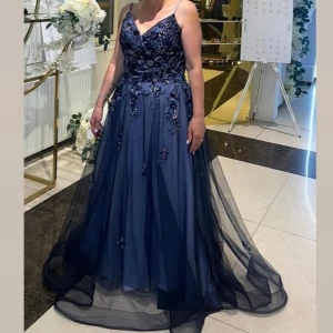 Marinblå långklänning med blommiga detaljer - Elegant marinblå långklänning med blommiga applikationer. Klänningen har en ärmlös design och ett lager av transparent tyg som ger en flytande effekt.