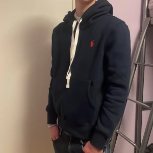 Mörkblå hoodie från Ralph Lauren - Säljer en snygg mörkblå hoodie från Ralph Lauren med det klassiska röda logotypen på bröstet. Tröjan har en ficka vid magen och vita snören vid huvan. Perfekt för en avslappnad stil. Tröjan är helt ny och aldrig använd så lappen finns kvar.