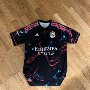 Svart Real Madrid fotbollströja från Adidas - Säljer en svart Real Madrid fotbollströja från Adidas med coolt mönster i rosa och blått. Tröjan har korta ärmar och klubbens emblem på bröstet. Perfekt för fans av laget! Riktig snygg och fräsch tröja inför sommaren den är för både män och kvinnor tröjan