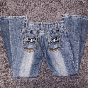 Blå jeans med rosett-detaljer från LJJ Jeans - Snygga blå jeans från LJJ Jeans med unika rosett-detaljer på bakfickorna. Jeansen har en klassisk femficksdesign och dekorativa knappar vid midjan. Byxorna är utsydda till bootcut därmed en extra söm, innerbenslängden 78 cm och tvärs lver midjan 37 cm ❤️ PRISET ÄR DISKUTERBART!!! ❤️