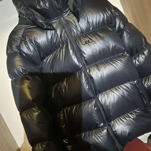 Svart pufferjacka från J.Lindeberg - Snygg svart pufferjacka från J.Lindeberg med dragkedja och huva. Perfekt för kalla dagar med sin quiltade design som håller värmen. Jackan har en stilren look och är ett måste för vintern.