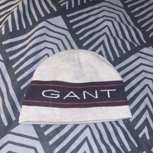 Grå mössa från GANT - Snygg grå mössa från GANT med ett brett svart band och röd rand. GANT-loggan är tydligt broderad i vitt framtill. Perfekt för kyliga dagar.
