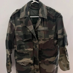Camouflage militärjacka - Säljer en cool militärinspirerad jacka i camouflage med knappar och flera fickor. Jackan har en klassisk krage och axelklaffar för en autentisk look. Perfekt för en avslappnad stil.