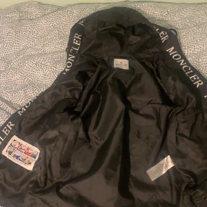 Moncler windbreaker  - Snygg svart jacka från Moncler med huva och dragkedja. Jackan har Moncler-logotypen på ärmen och text längs huvan. Perfekt för kyligare dagar. En moncler windbreaker 