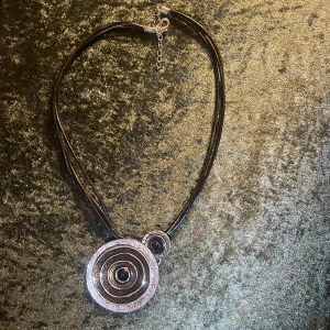 Svart halsband med cirkulärt hänge - Unikt svart halsband med ett stort cirkulärt hänge i silverfärg. Hänget har en detaljerad design med flera lager och en central pärla. Halsbandet har en justerbar kedja och stängs med en karbinhake.
