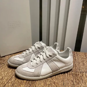 Vita sneakers från Maison Margiela - Snygga vita sneakers från Maison Margiela med gråa detaljer. Skorna har en klassisk design med snörning och är tillverkade i en kombination av skinn och mocka. Skorna är självklart äkta! Box och lite mer saker ingår. På köpet ingår även en vit  ami Paris T-shirt!!