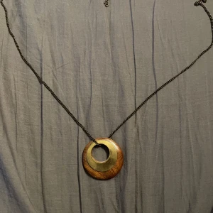 Halsband med cirkulär hängsmycke i trä och metall - Unikt halsband med ett stort cirkulärt hängsmycke i trä och metall. Kedjan är mörk och smycket har en stilren design med en kombination av naturligt trä och metallisk yta. Perfekt för att ge en bohemisk touch till din stil.
