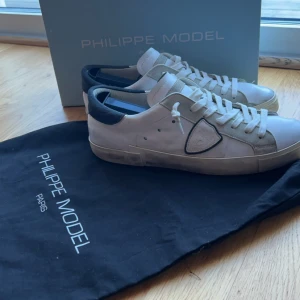 Vita sneakers från Philippe Model - Snygga vita sneakers från Philippe Model med svarta detaljer och snörning. Skorna har en klassisk design med en diskret logotyp på sidan. Perfekta för en stilren look. skorna är i jättebra condition och har endast använts i en månad. Köpte dem på kicksnstuff. målat med en skofärg på ställen där det fanns repor men det märks inte överhuvudtaget och det ser ändå bra ut under. Inget OG