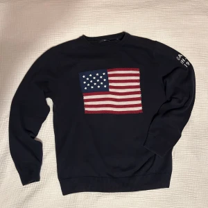 Svart tröja med amerikansk flagga från Gant - Snygg svart tröja från Gant med en stor amerikansk flagga på framsidan. Tröjan har långa ärmar och texten 'GANT NH-CT 1949' på ärmen. 