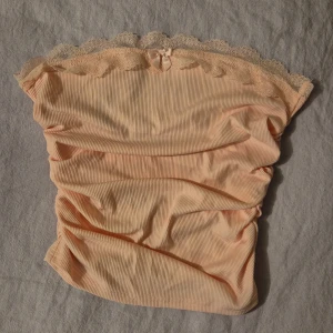 Beige ribbad tubtopp med spetsdetaljer - Säljer en superfin babyrosa ribbad tubtopp med vacker spetskant runtom.. Toppen har en söt liten rosett framtill som ger den en extra touch. Perfekt för sommardagar eller en festlig kväll!