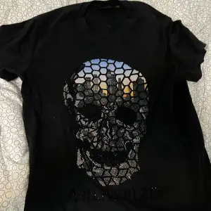 Säljer en cool svart t-shirt från Philipp Plein med ett stort dödskallemotiv på framsidan. T-shirten är kortärmad och gjord av 100% bomull, vilket gör den superbekväm. Perfekt för en avslappnad stil med en edgy touch!